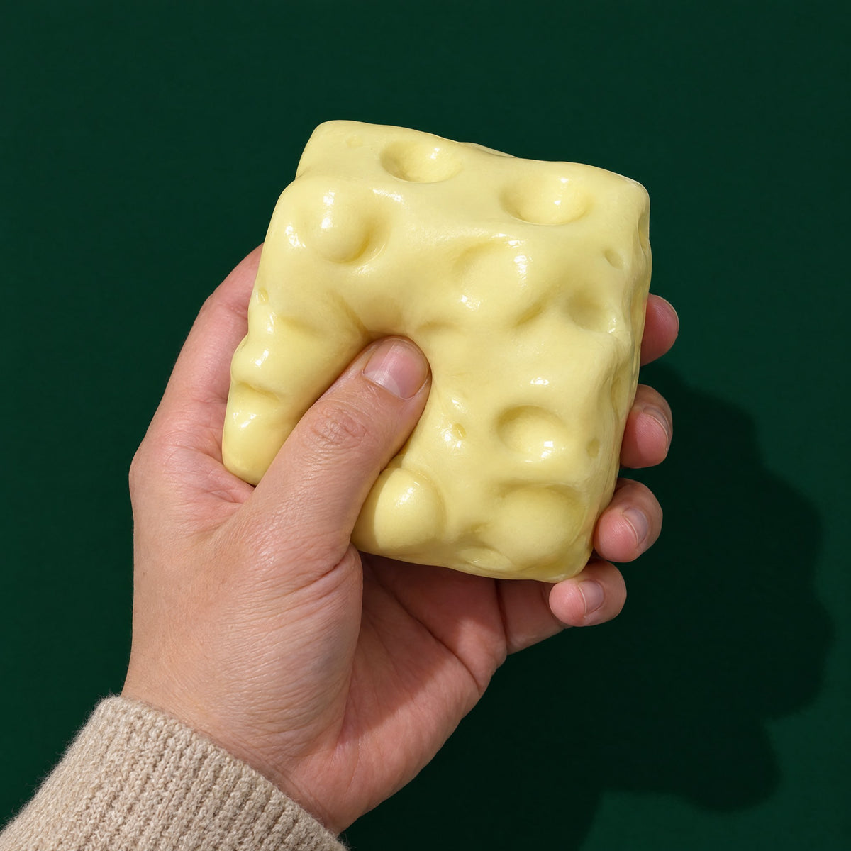 Adult thumb pressing deep into a pale yellow Swiss-cheese squishy cube, showing how soft it is