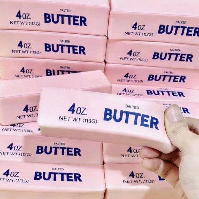 Real photo: a hand squeezing a pink Crunch Butter block so it bends, in front of stacked pink blocks