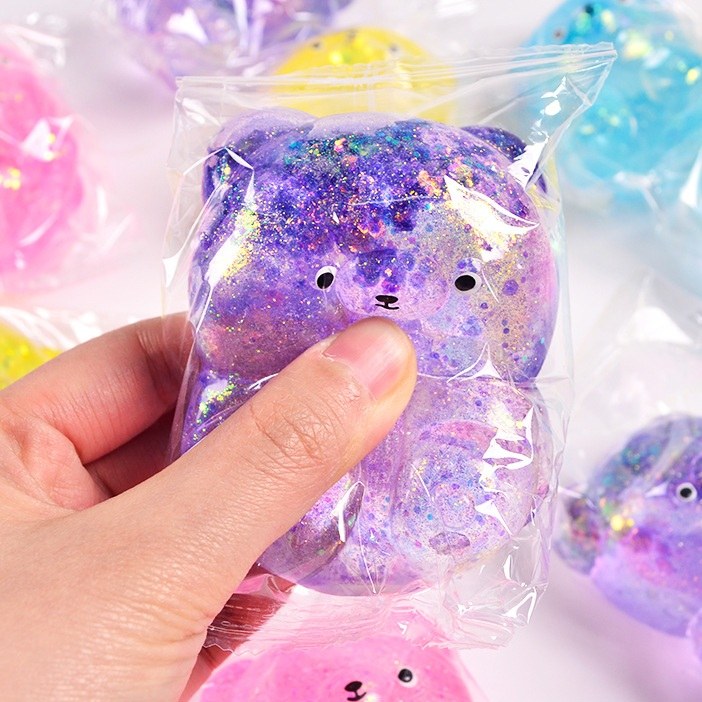 Real photo: purple glitter-gel bear squishy sealed in its clear plastic bag, held in an adult hand
